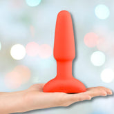 A hand holds the B-Vibe Rimming Plug 2 Vibrating Butt Plug in orange by B-Vibe, featuring a wide base and tapered tip, designed for pleasurable rimming sensations, set against a softly blurred pastel bokeh background.